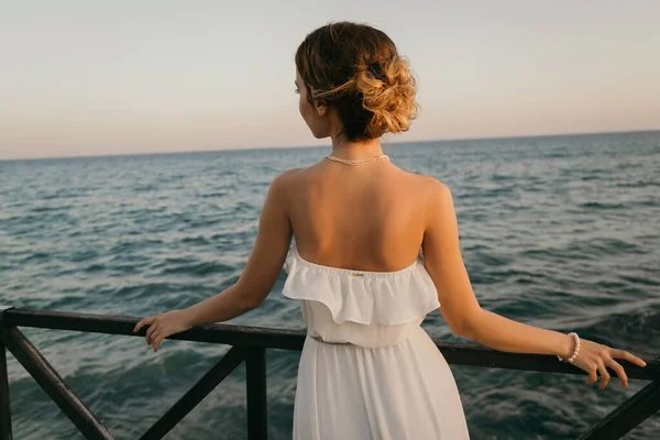 ギリシャ風の白いドレスを着た美しい若い笑顔の女の子が海の遊歩道を歩いています。夏時間日没時間。海での花嫁結婚式 — ストック写真