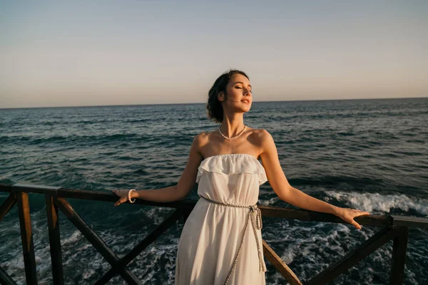 Beyaz elbiseli, Yunan usulü gülümseyen güzel bir kız deniz kenarında yürüyor. Yaz, gün batımı. Denizde gelin düğünü 