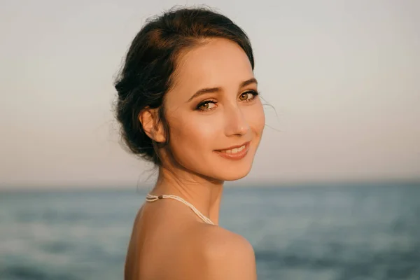 Beyaz elbiseli, Yunan usulü gülümseyen güzel bir kızın portresi deniz kenarında yürüyor. Yaz, gün batımı. Gelin, denizde düğün 