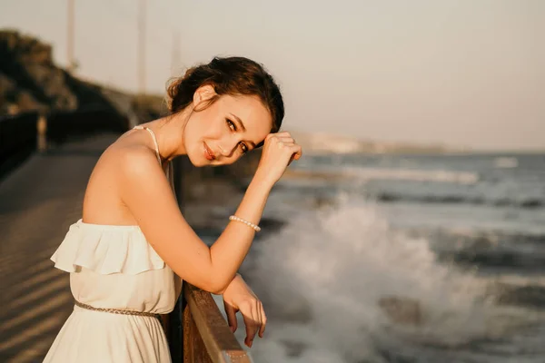 Portrét krásné mladé usměvavé dívky v bílých šatech v řeckém stylu kráčející podél mořské promenády. Léto, západ slunce. Nevěsta, svatba na moři — Stock fotografie