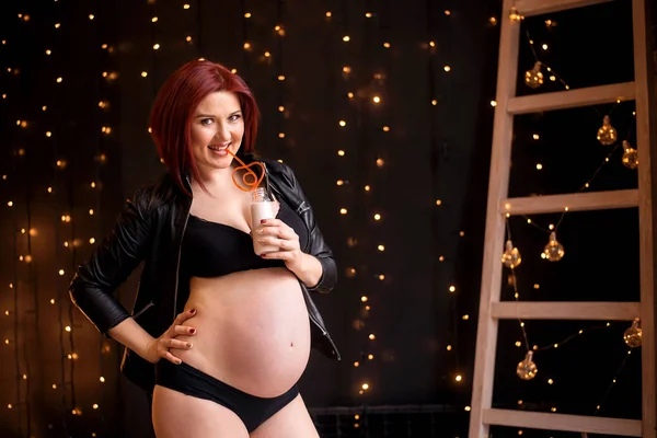 Beautiful audacious pregnant woman at 26 weeks in lingerie and jacket standing and drinking milk from bottle with neon straw