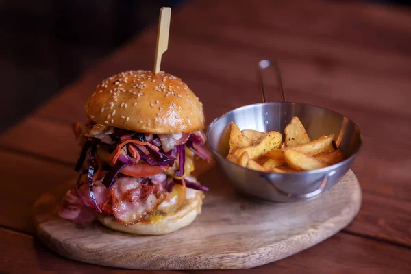 Big burger with red onion, tomato, cheese, beef and bacon meat served with fried potatoes on wooden plate in bar or cafe