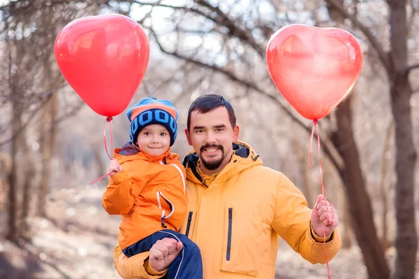 Mother\'s day and International woman\'s day concept: father and toddler son standing outdoors with balloons shaped like hearts and smiling. Leafless trees in background