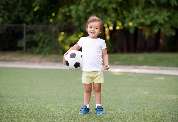 小さなサッカー選手の肖像:ボールと笑顔でサッカー場に立ってスポーツの制服を着た幸せな幼児の少年。活動的な子供時代のコンセプト — ストック写真