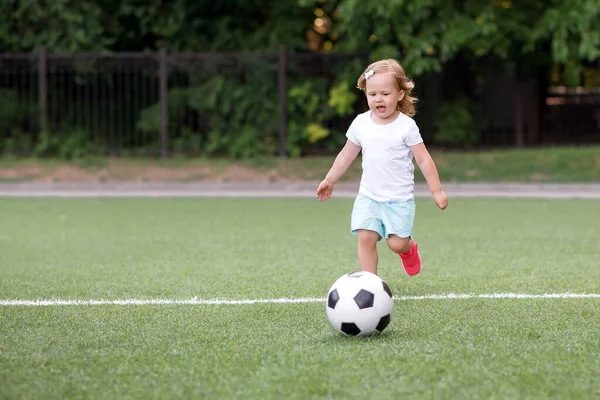 幼児の女の子サッカーをプレイ:ブロンドの子供がサッカーボールをキックするスタジアムで実行されます。屋外活動、スポーツゲーム、幸せな子供時代のコンセプト — ストック写真