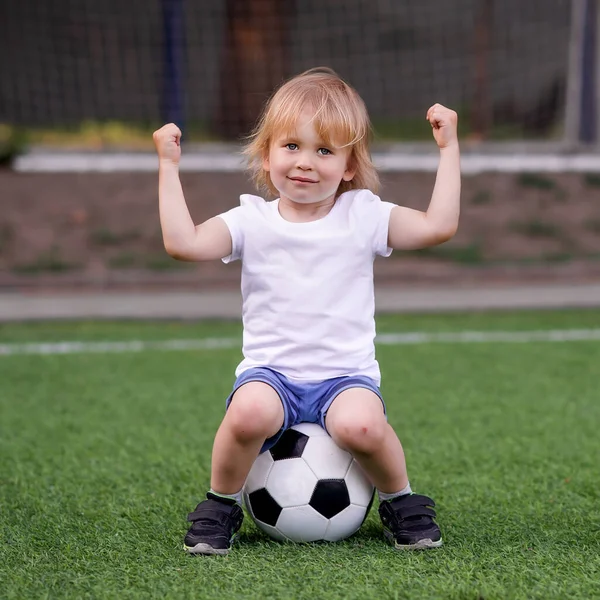 小さなゴールキーパー ブロンドの幼児の子供が腕を上げてサッカーボールの上に座って 冗談で筋肉と力を示しています 幼稚園のコンセプトで成功 ジムクラス — ストック写真