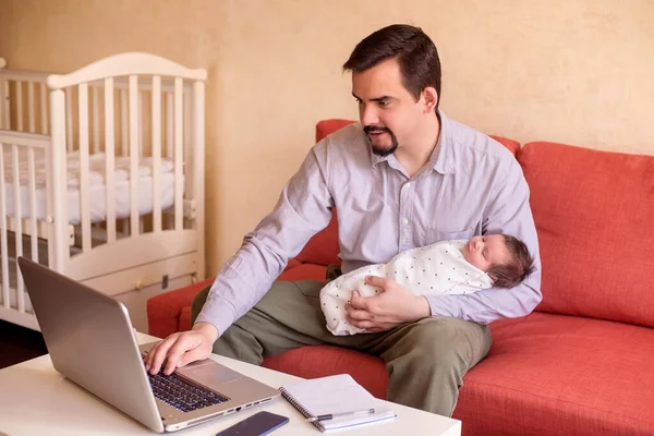 リモートワーカー グレーのシャツを着た若い父親がソファに座って 片手で幼児の赤ちゃんを抱えて 同時にノートパソコンのキーボードを入力します 小さな子供の概念を持つ家族の男性のための家からの仕事や管理 ストック画像