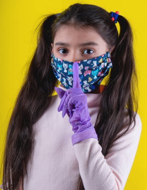 Tek kullanımlık eldivenleri ve sarı arka planda renkli maskesi olan tatlı bir kız. Covid-19 'u durdurun. Evde kal. Konsept. Koruyucu tıbbi maskeli genç bir kız. Karantinaya alın. Coronavirus. Kişisel tecrit.