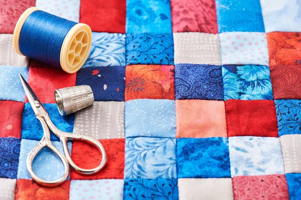 Schaar, draad en een vingerhoed liggend op het blauwe en rode vierkante stukken stof aan elkaar genaaid — Stockfoto