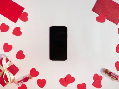 Ortasında cep telefonu olan kompozisyon, beyaz yüzeyde hediyeler ve kırmızı kalpler. Üst Manzara. Metin için boşluk