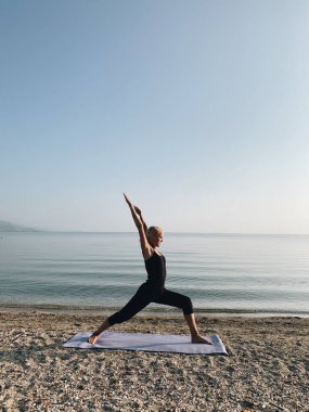 Sarışın kız gün doğumunda deniz kıyısında savaşçı yogası yapıyor.