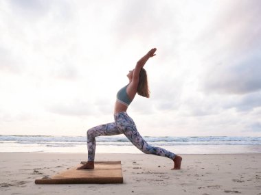 Genç beyaz kadın, gün doğumunda deniz kıyısında yoga savaşçısı pozu veriyor..