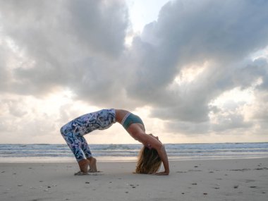 Genç esnek kız günbatımında sahilde tekerlek yogası pozu.