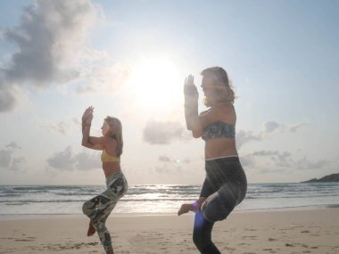 İki kadın güneş ışığında deniz sahilinde kartal yogası yapıyor.