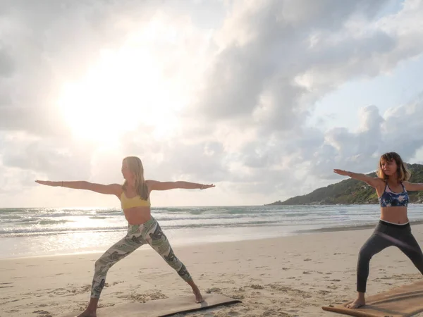 Two women practice warrior yoga posture at sunset seashore