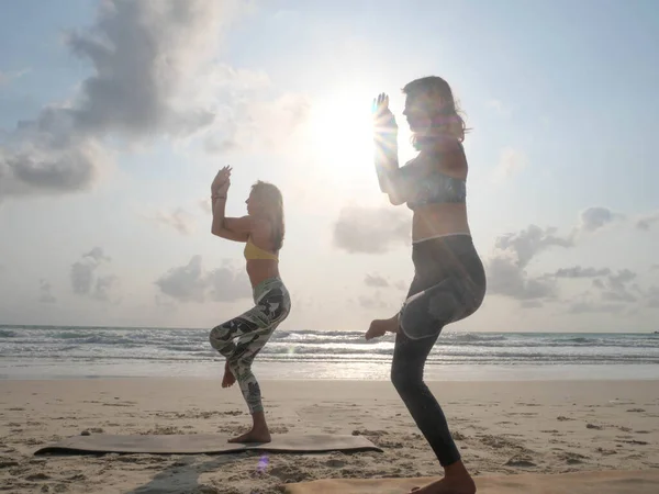Two women practice eagle yoga asana at sea beach in sunshine