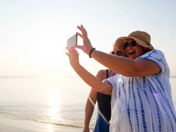 Dos anciana divertida haciendo selfie en el teléfono inteligente al atardecer playa — Foto de Stock