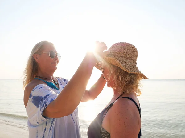 Feliz mulher sênior colocando um chapéu em sua namorada lá fora na praia — Fotografia de Stock