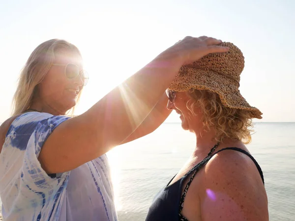 Mulher sênior colocando um chapéu em sua namorada lá fora na praia à luz do sol — Fotografia de Stock