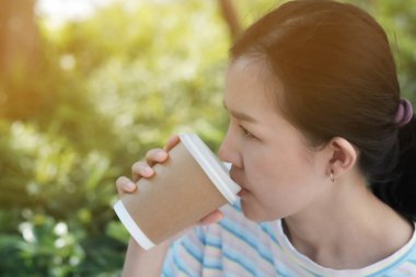 Asyalı bir kız kahve içiyor. Bahçedeki kafede, kağıt kahve bardağının içinde. 