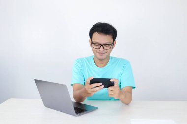 Asyalı genç adam iş yerinde oyun oynarken akıllı telefondan zevk alır. Endonezyalı adam mavi gömlek giyiyor..