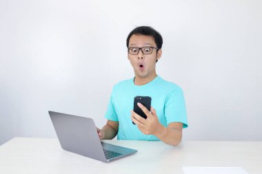 Genç Asyalı adamın yüzü yanında dizüstü bilgisayarla telefonda gördüklerini şok etti. Endonezyalı adam mavi gömlek giyiyor..