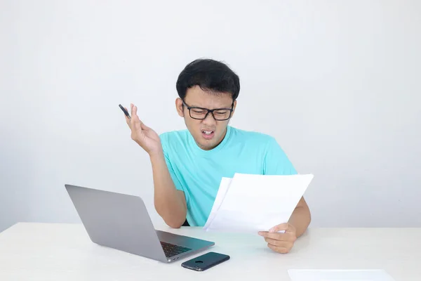 Wow face of Young Asian man shocked what he see at the document and laptop when working isolated grey background. Indonesian man wearing blue shirt.