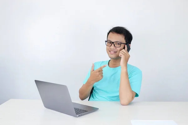 幸せな顔をした若いアジア人男性は テーブルの上にラップトップを持つ携帯電話で話しています 青いシャツを着たインドネシア人男性 — ストック写真