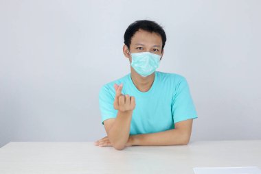 Hastalıktan korunmak için maske takan genç bir Asyalı Koreli, izole beyaz arka planda aşk işareti gösteriyor. Coronavirus konsepti. Endonezyalı adam mavi gömlek giyiyor..
