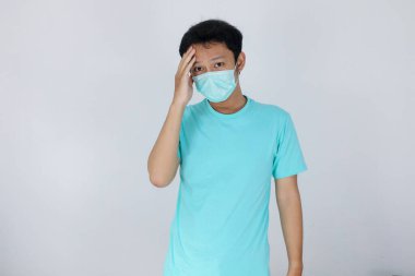 Tıbbi maske takan genç Asyalı adam şiddetli baş ağrısından muzdarip, parmaklarını şakaklarına bastırıyor, çaresiz yüz ifadesiyle acıyı dindirmek için gözlerini kapatıyor..