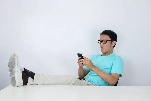 Young Asian Man with happy face with what he see on a mobile phone with leg on the table. Indonesian man wearing blue shirt.