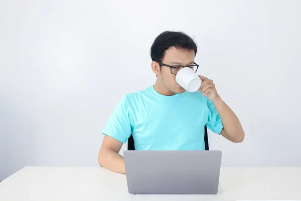 白ガラスのドリンクコーヒーでノートパソコンで働く若いアジア人男性は本気です インドネシア男は青いシャツを着て隔離された灰色の背景 — ストック写真