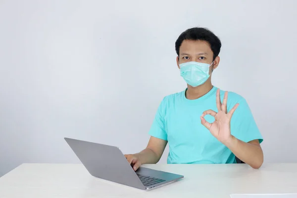医療用マスクをした若いアジア人男性は テーブルの上のラップトップで作業するときに深刻であり 焦点です 青いシャツを着たインドネシア人男性 — ストック写真