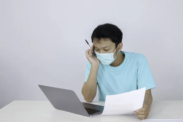 医療用マスクを身に着けている若いアジアの男は不健康な感じです 疲れています テーブルの上にドキュメントやノートパソコンで作業と混同 — ストック写真