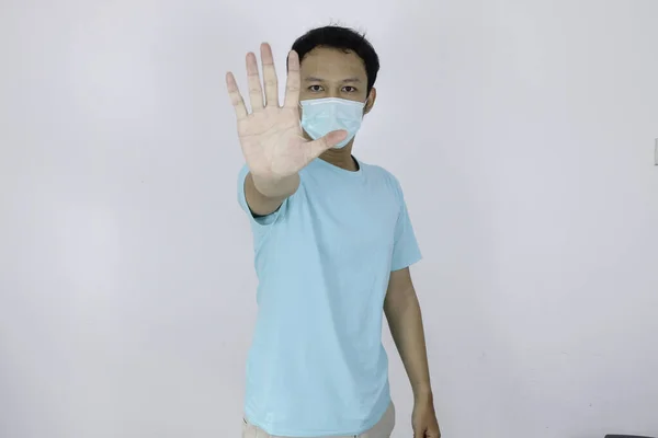 若いアジアの男は 衛生的なマスクを着用するか ジェスチャーを停止手でパンクしている コロナウイルス感染やインフルエンザなどの呼吸器疾患の恐れがあります 白地に隔離されたインドネシア人男性 — ストック写真