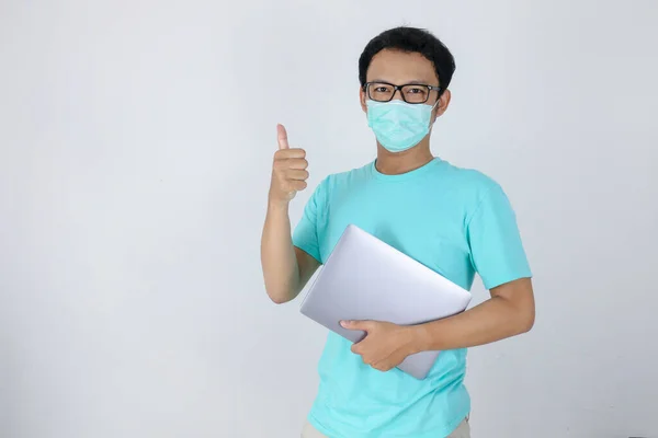 若いアジアの男は 医療用マスクを着用し Okの手のサインをノートパソコンを運んでいる 青いシャツを着たインドネシア人男性 — ストック写真
