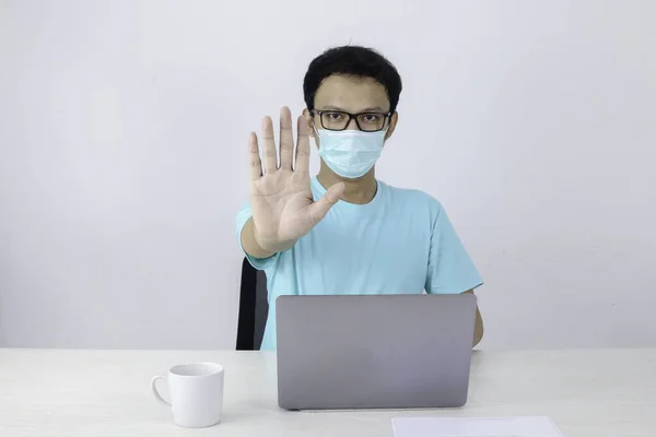 Genç Asyalı adam hijyenik maske takıyor ya korkuyor ya da dizüstü bilgisayarda çalışırken el hareketi yapıp panikliyor. Koronavirüs enfeksiyonundan ya da grip gibi solunum hastalıklarından korkuyor..