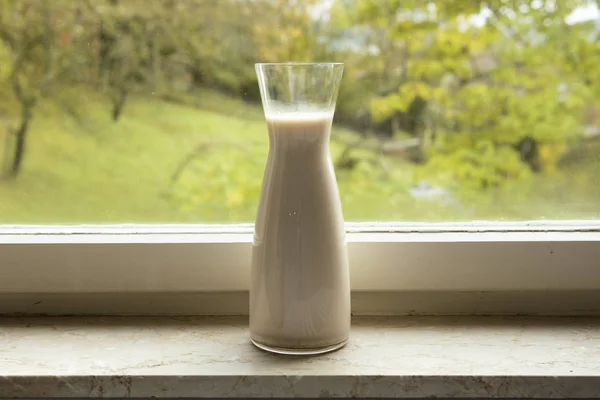 Homemade plant milk in glass bottle standing on the window sill. Cozy atmosphere in kitchen .Green window background. Dairy free diet, healthy lifestyle.