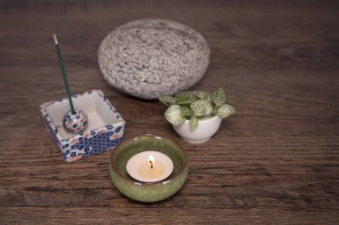 Wellnes arrangement. Burning candle in a ceramic pot, incense sticks, stone and fresh plants on wooden background. Zen style spirituality decoration. Daily meditation routine.