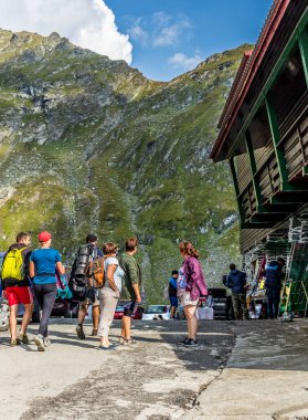 Bir dağ dağ evinin yan manzarası, dağ yolunda yürüyen insanlar, arka planda dağlar ve yukarıda mavi bir gökyüzünde kabarık bulutlar.