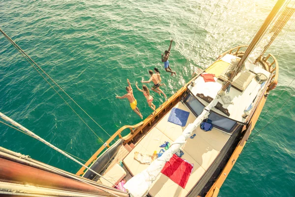 Vista aérea de los jóvenes que saltan desde el barco de vela en viaje de mar - Ricos amigos felices divirtiéndose en el día de fiesta de verano - Concepto de vacaciones exclusivas - Filtro vintage cálido con halos de destellos solares mejorados —  Fotos de Stock