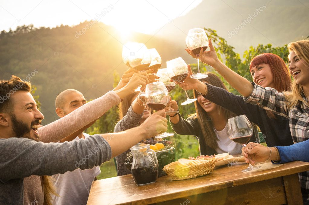 Happy friends having fun outdoors - Young people enjoying harvest time together at farmhouse vineyard countryside - Youth and friendship concept - Focus on hands toasting wine glasses with sun flare