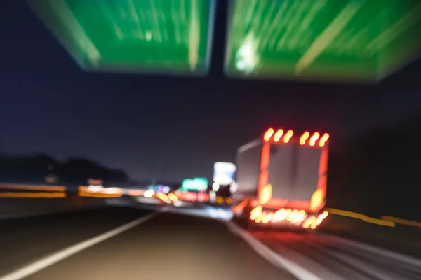 Defocused blurred motion of semi truck speeding on highway under street signs - Night traffic and transport logistic concept with semitruck container driving on speedway - Bokeh and tilted composition — Stock Photo, Image