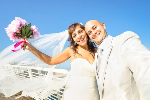 Young newlyweds with bouquet taking selfie against blue sky - Wedding couple cheering together after ceremony - Lovely life concept with groom and bride celebrating love forever on a warm sunny day — Stock Photo, Image