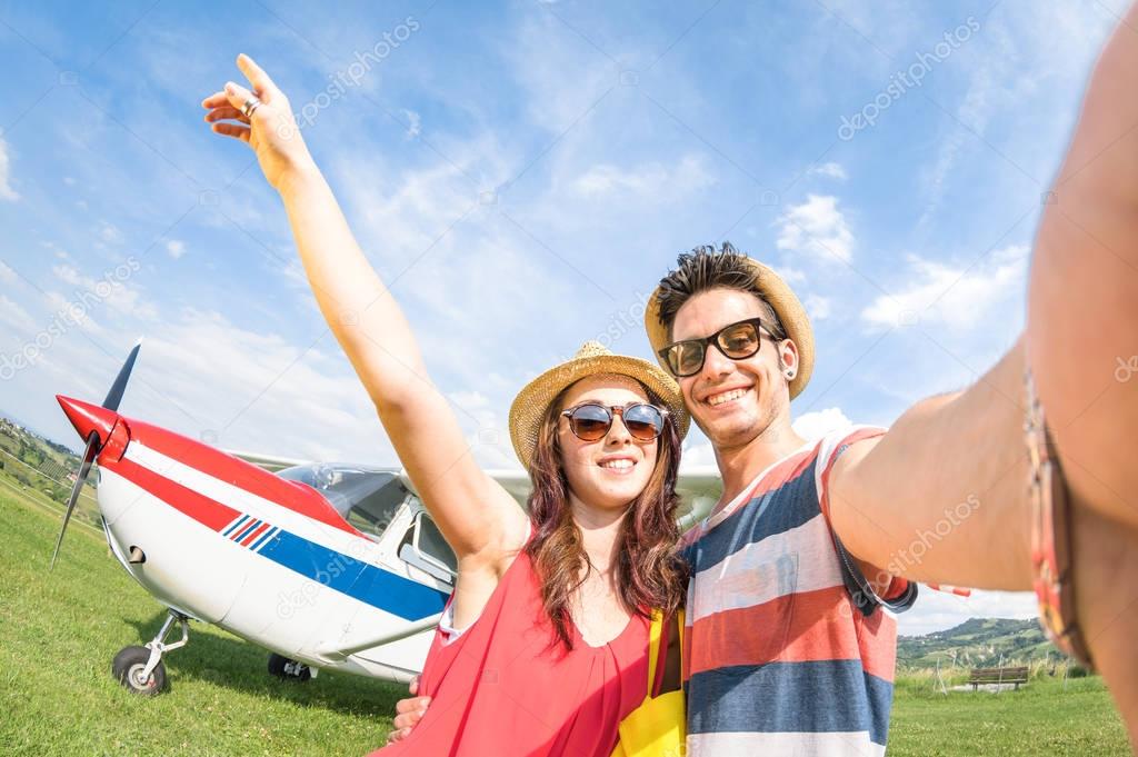 Young couple taking selfie with lightweight airplane - Happy people boarding on excursion air plane - Alternative adventure vacation concept - Warm day colors with tilted horizon fisheye distortion