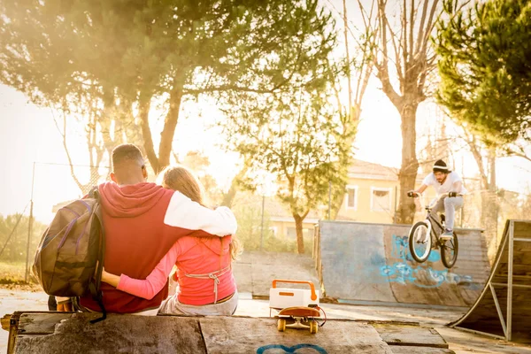 Casal multirracial apaixonado sentado no parque de skate com música assistindo amigos na exposição bmx freestyle - Conceito de relacionamento urbano com os jovens se divertindo ao ar livre - Filtro contrastado quente — Fotografia de Stock