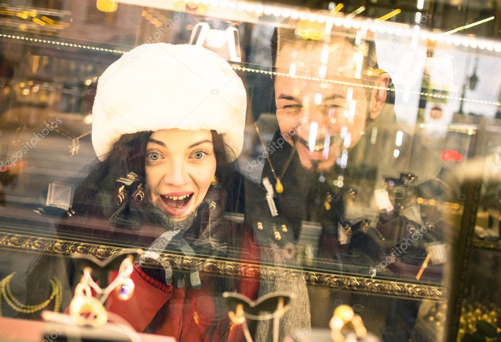 Modern hipster couple shopping on winter cloth pointing jewelry store window display - Consumerism concept with young fashion people looking for Christmas present at urban city tour - Focus on woman