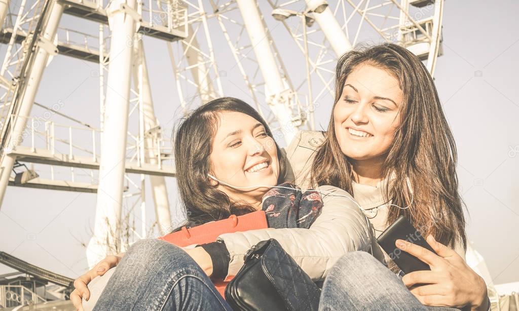 Best friends enjoying time together - Young people with smartphone music playlist in autumn sunny day - New trends and technology concept with hipster girlfriends having fun outdoors at ferris wheel