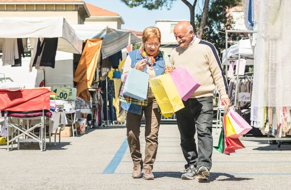 Casal sênior fazendo compras juntos no mercado de pulgas com a esposa assistindo em sacos de marido - Conceito de idoso ativo com homem maduro e mulher se divertindo na cidade - Happy retired people moments on vivid colors — Fotografia de Stock