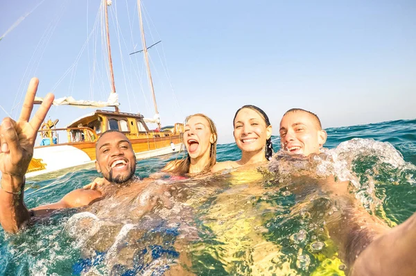 Jóvenes amigos multirraciales tomando selfie y nadando en un viaje en barco de vela - Chicos y chicas felices ricos divirtiéndose en el día de la fiesta de verano - Concepto de vacaciones exclusivas - Filtro cálido de la tarde brillante —  Fotos de Stock